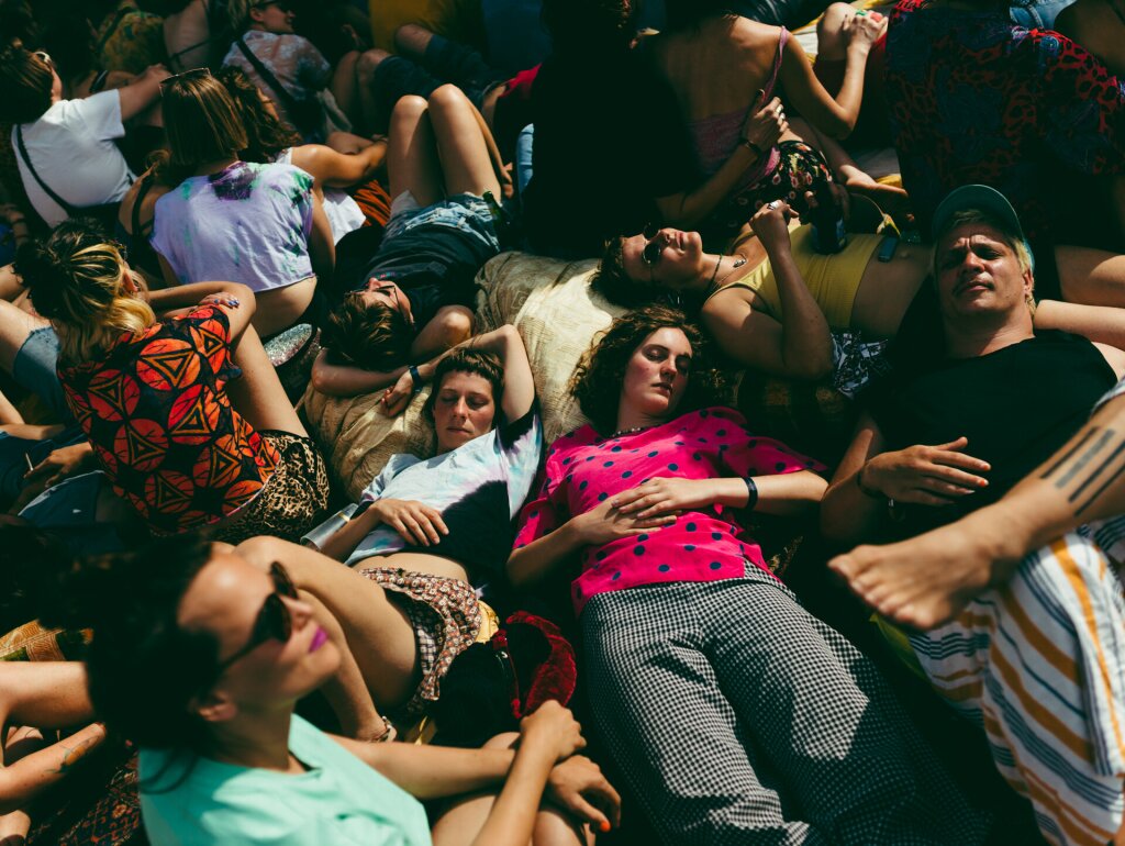 A group in colourful clothing lies relaxed on a few blankets, surrounded by lots of people doing the same.